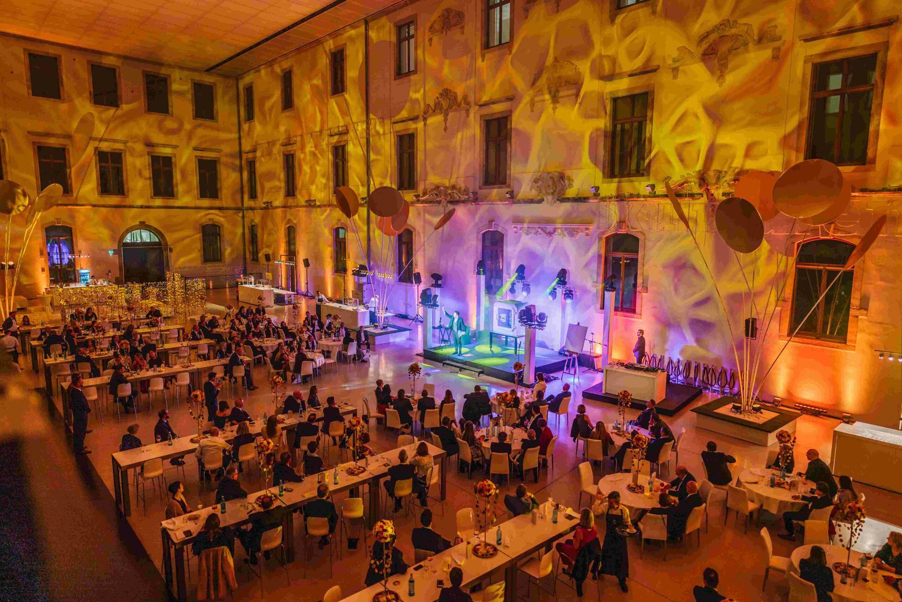 Gala dinner in a courtyard washed in warm golden light, guests seated facing a lit stage